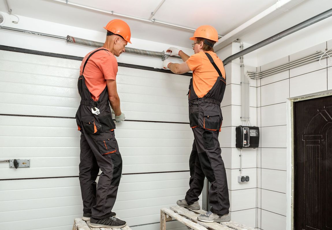 Two workers in orange shirts and dark overalls stand on step stools, installing a garage door track system indoors.