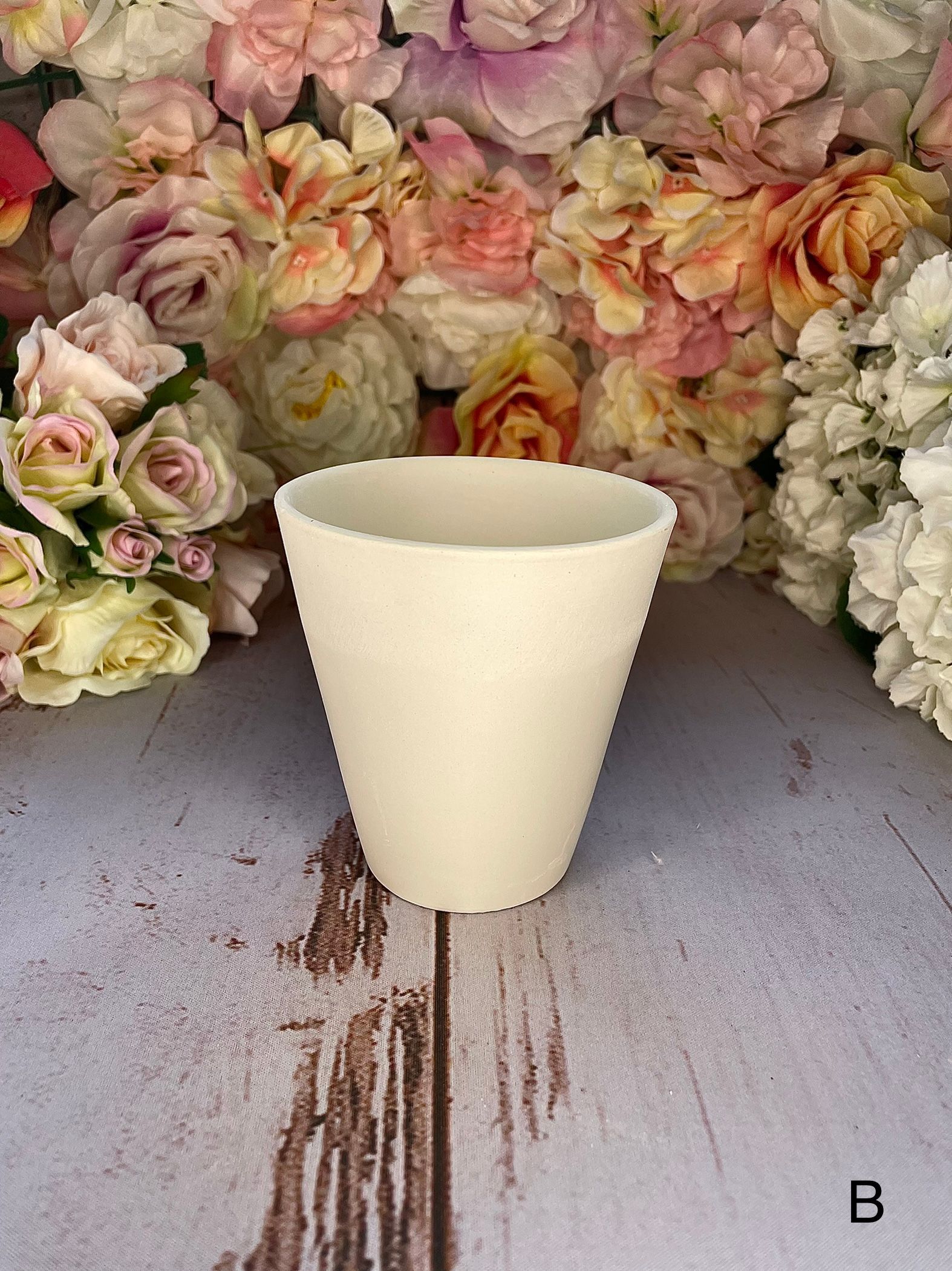 A white vase is sitting on a wooden table in front of a bunch of flowers.