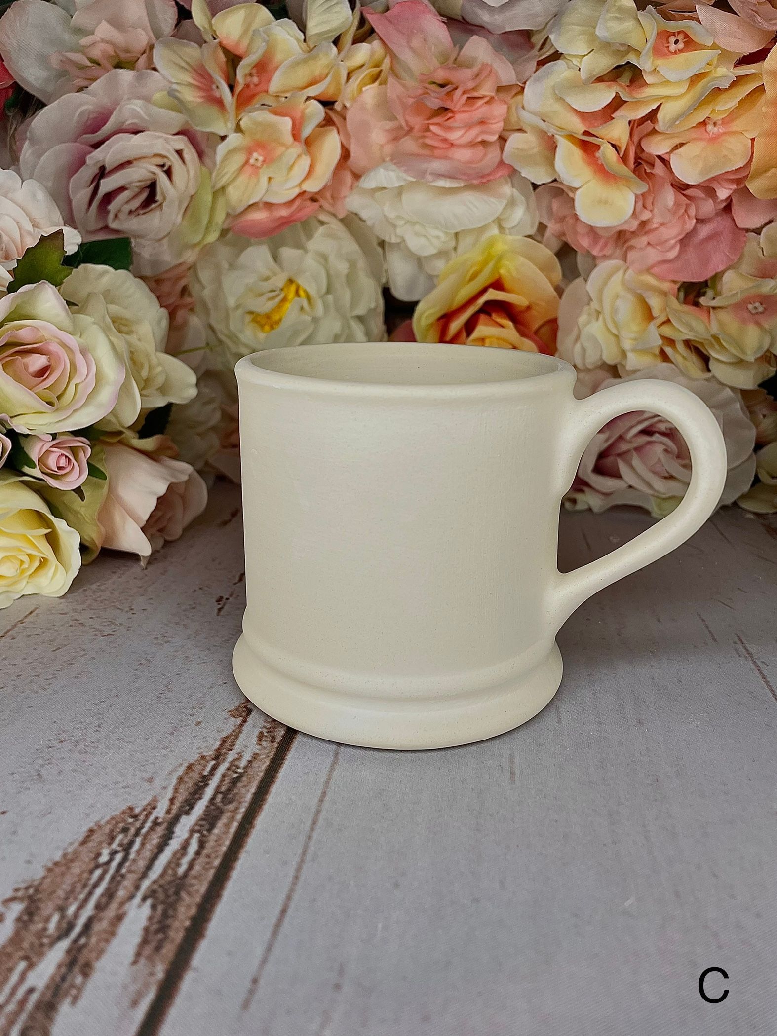 A white mug is sitting on a wooden table next to flowers.