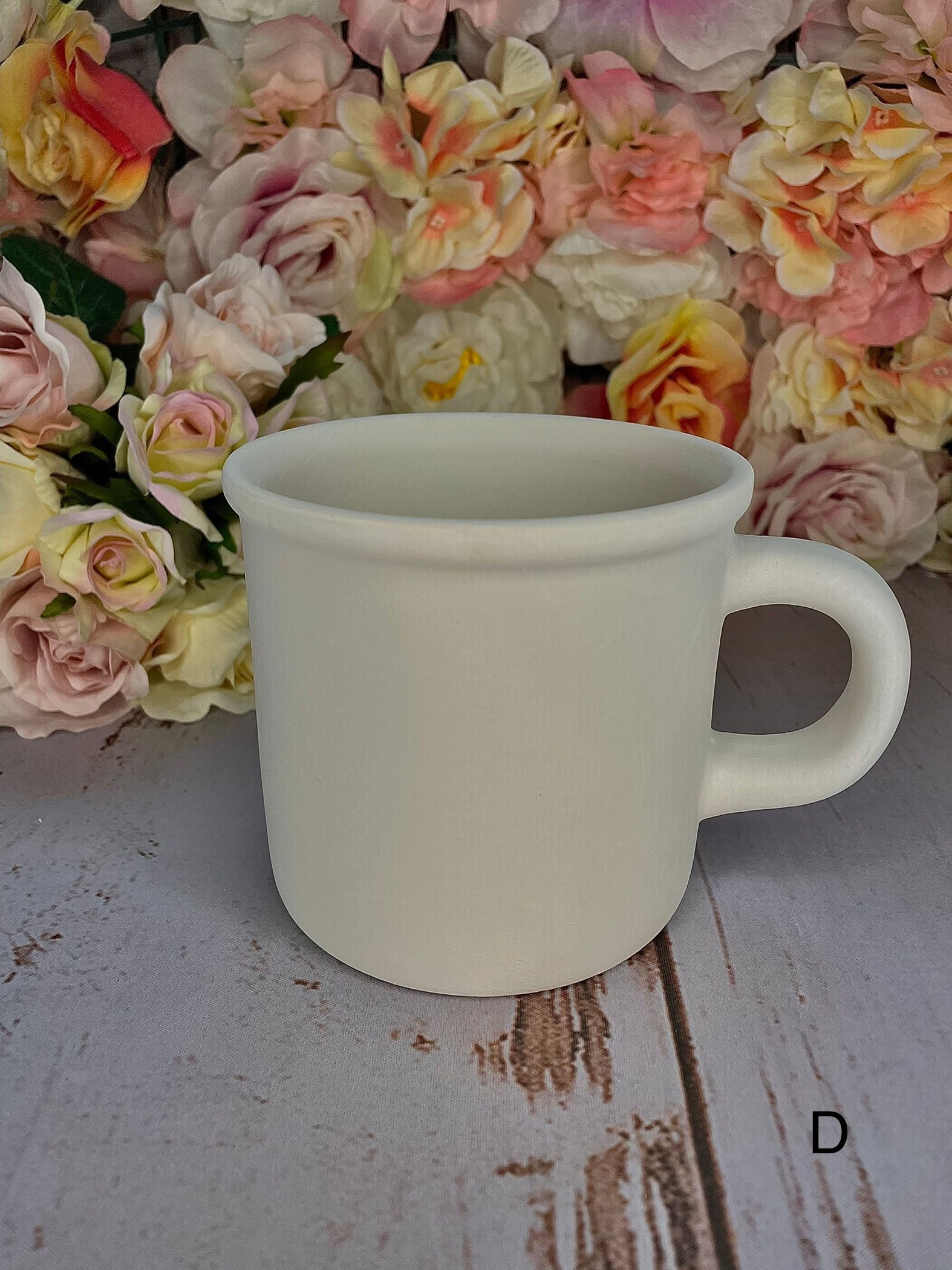 A white mug is sitting on a wooden table in front of a bouquet of flowers.