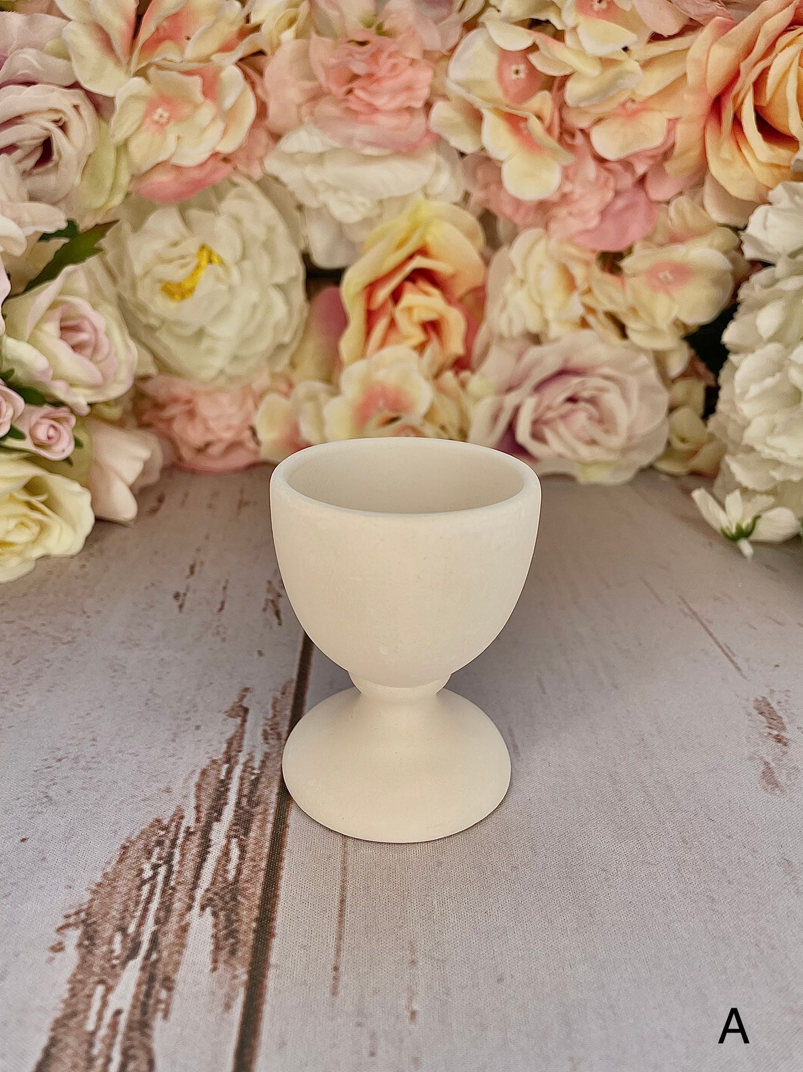 A white egg cup is sitting on a wooden table in front of flowers.