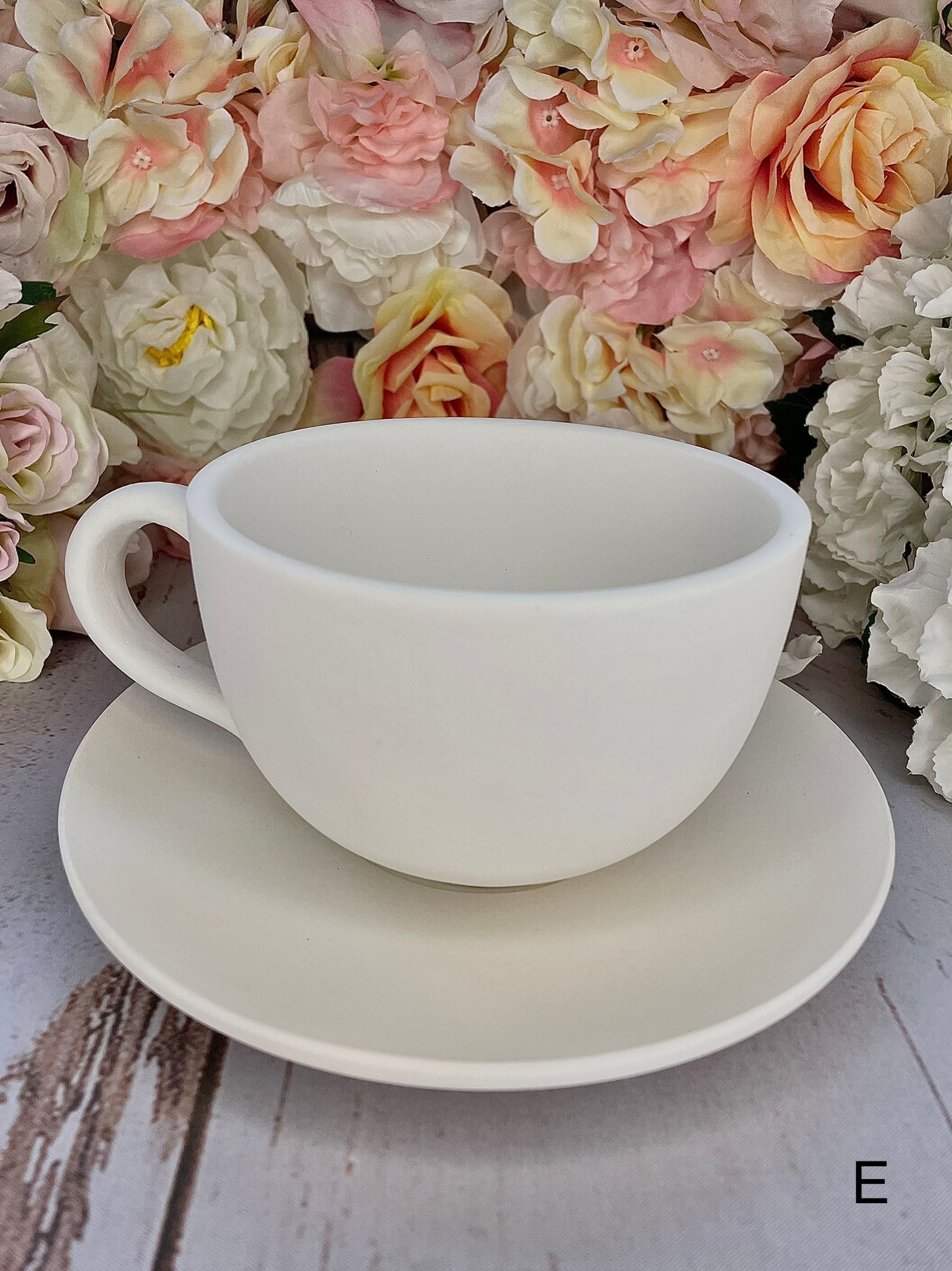 A white cup and saucer are sitting on a wooden table with flowers in the background.