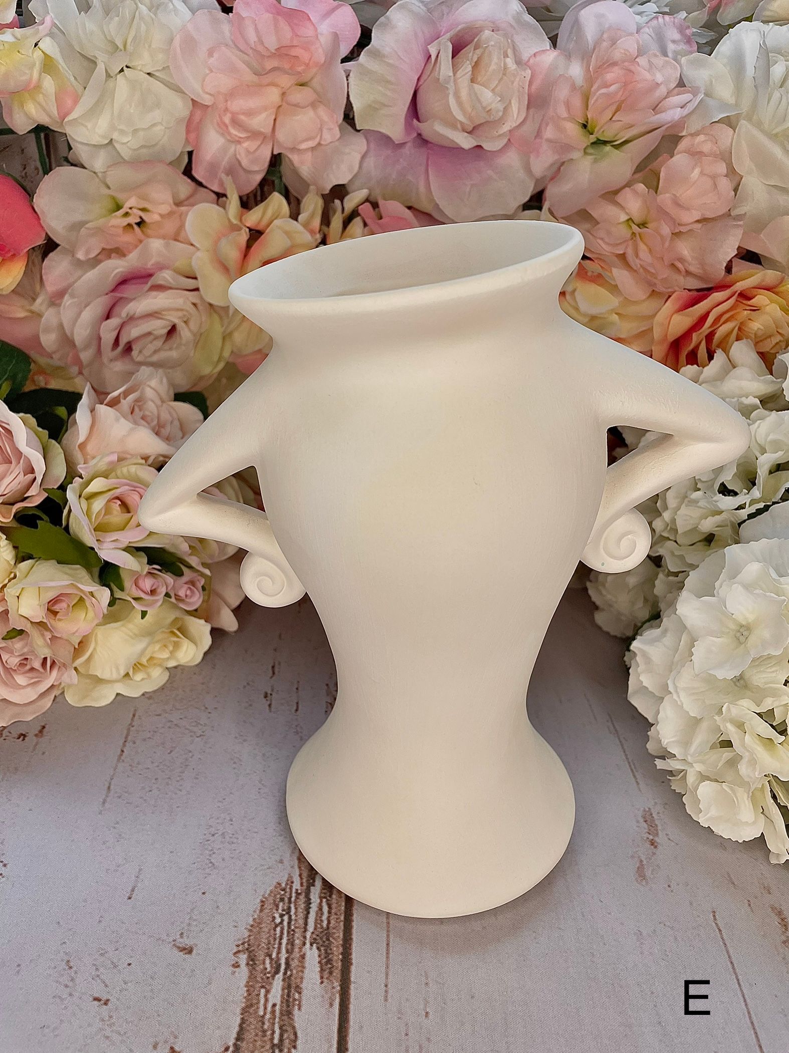 A white vase with two handles is sitting on a wooden table surrounded by flowers.