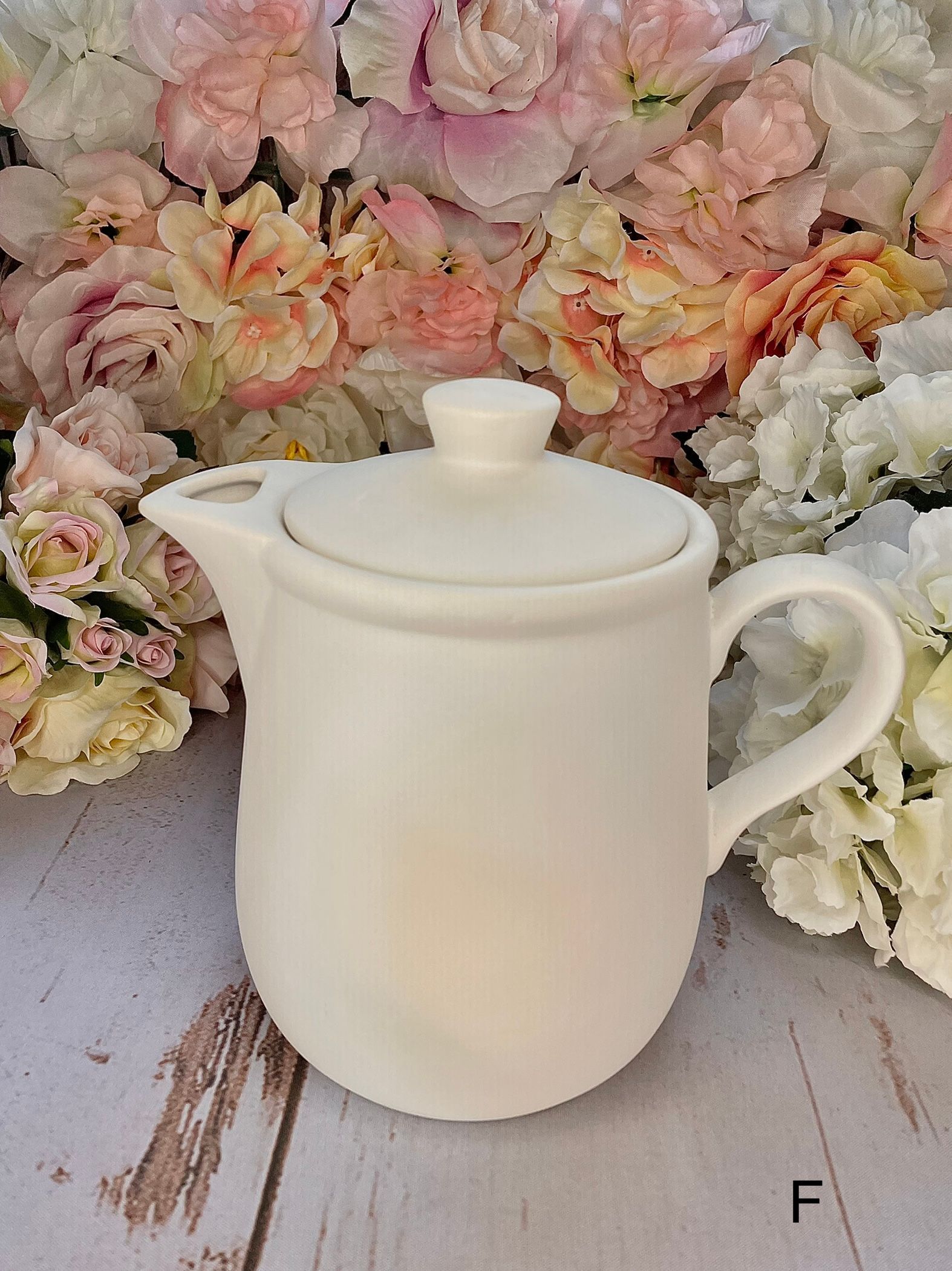 A white teapot is sitting on a wooden table next to flowers.