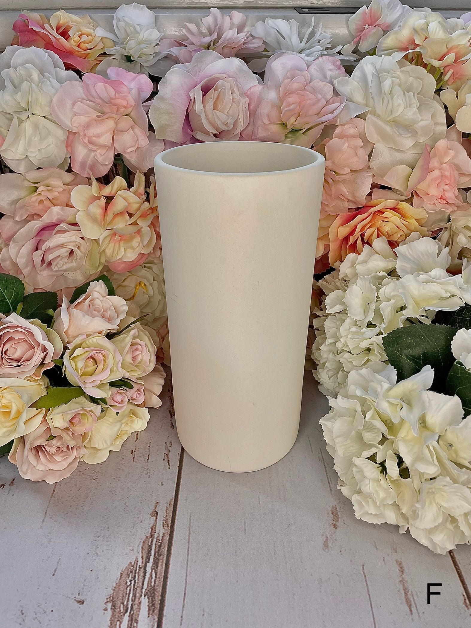 A white vase is sitting on a wooden table surrounded by flowers.