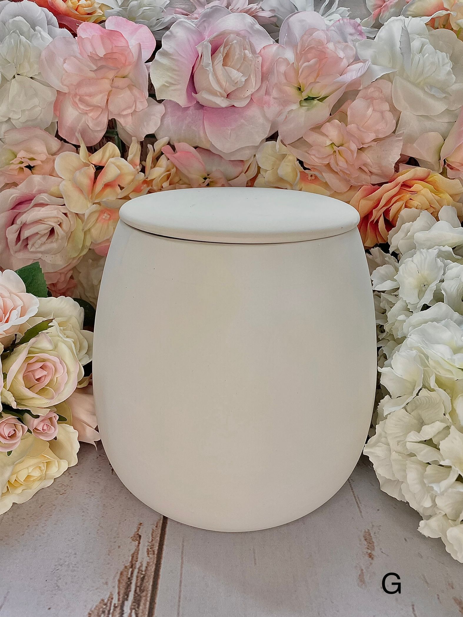 A white candle is sitting on a wooden table in front of a wall of flowers.