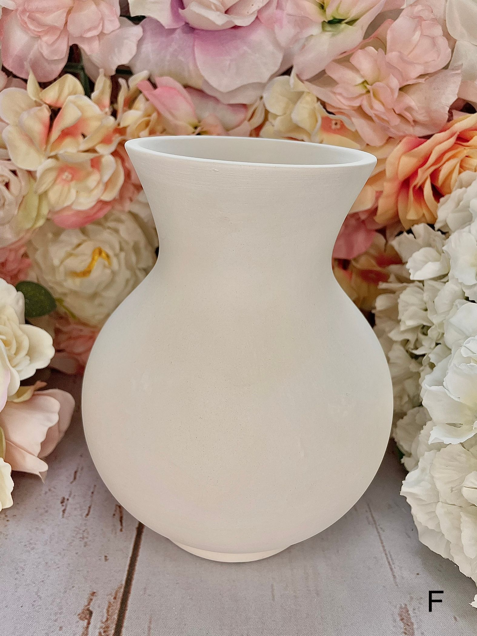 A white vase is sitting on a wooden table surrounded by flowers.