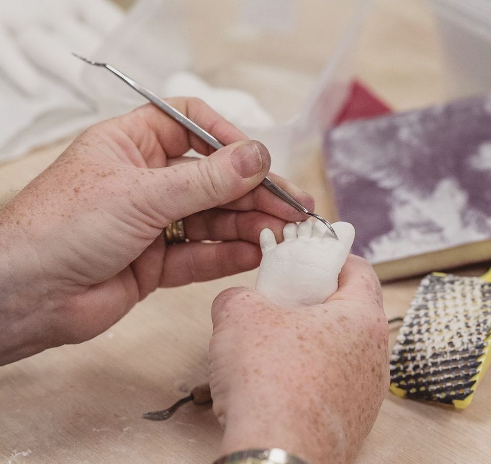 Hands sculpting a small, white, foot-shaped object with a metal tool on a wooden surface.