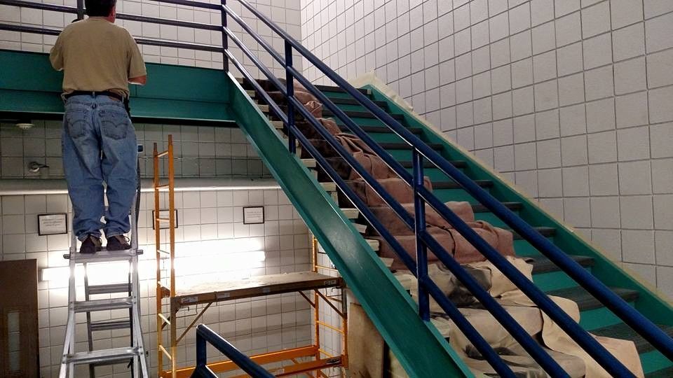 Person on a ladder painting near a turquoise staircase with brick steps. White tiled walls.