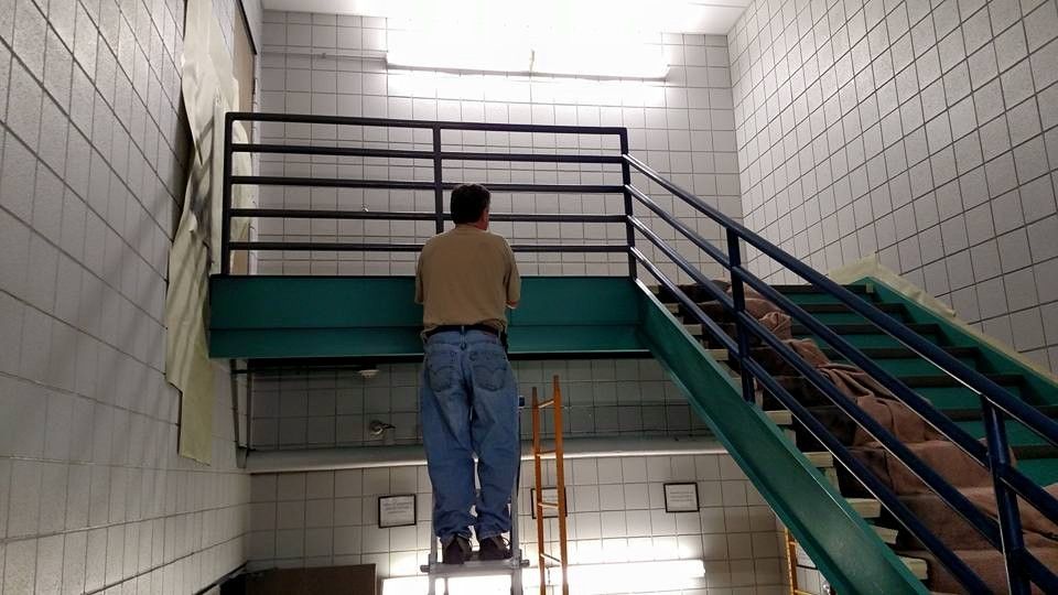 A person stands on a step ladder, working on a wall near a staircase with blue rails.