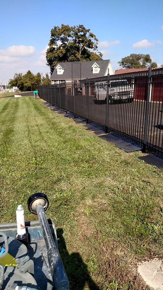 A long dark fence bordering a grassy area with a house in the background on a sunny day.