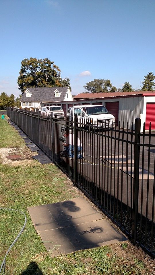 Black metal fence along a property, with a white van and a house in the background on a sunny day.