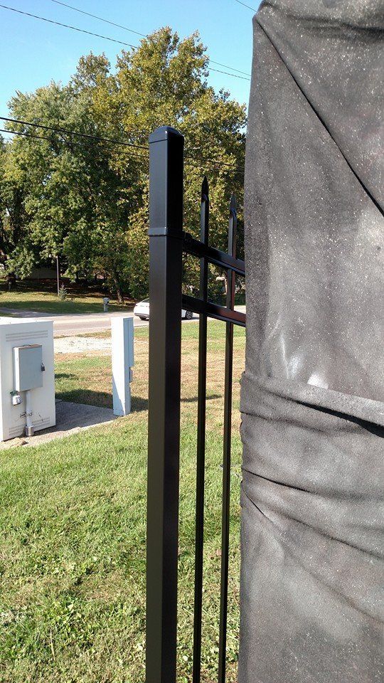 Black metal fence post next to a large, draped object. Green grass and trees in background.