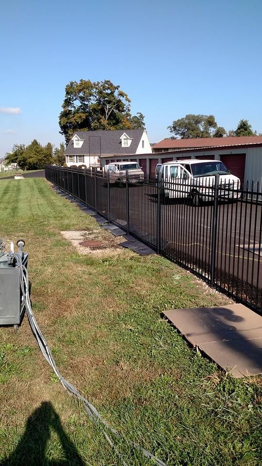 Black metal fence runs along a grassy area next to a white house and parked vehicles on a sunny day.