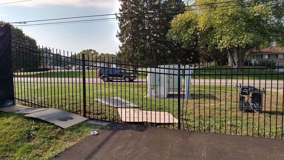 Black metal fence bordering a grassy area with a utility box and asphalt path. A vehicle is visible in the background.