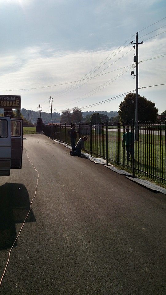 Three people installing a black fence along a paved road under a cloudy sky.
