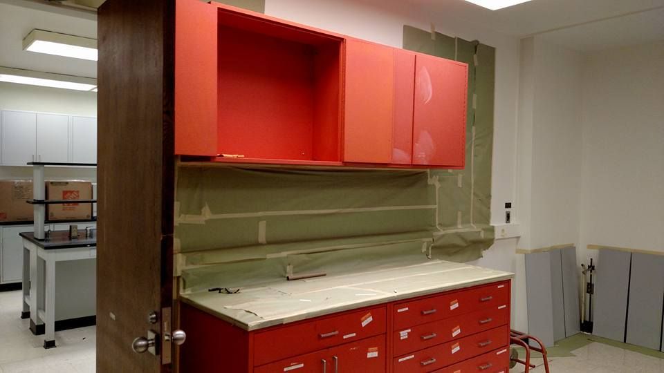 Red cabinets and drawers in a lab with opened upper cabinets and exposed wall.