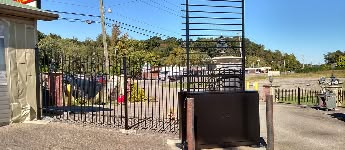 A black gate opens to a gravel lot with cars, trees, and buildings against a blue sky.