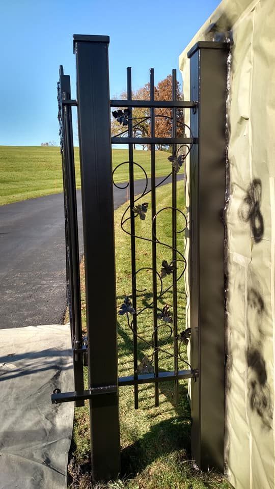 Black metal gate with decorative vines, attached to a stone column.  Green grass and blue sky in the background.