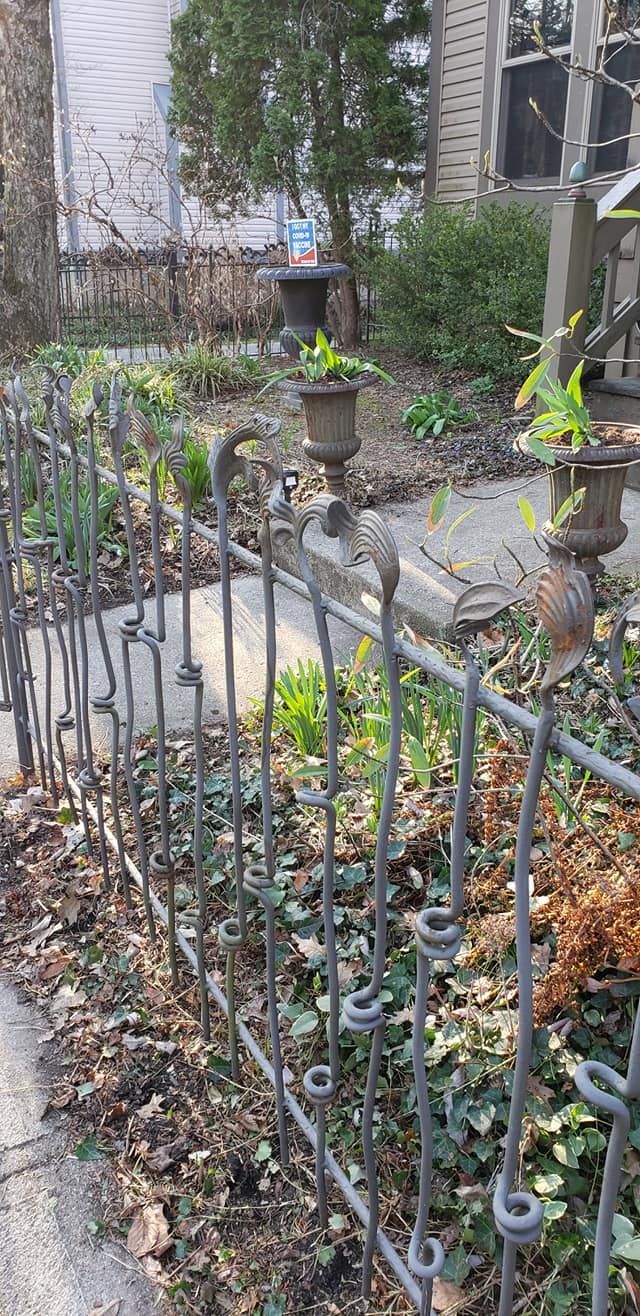 Wrought-iron fence with decorative swirls, bordering a garden bed with plants, urns, and a path.