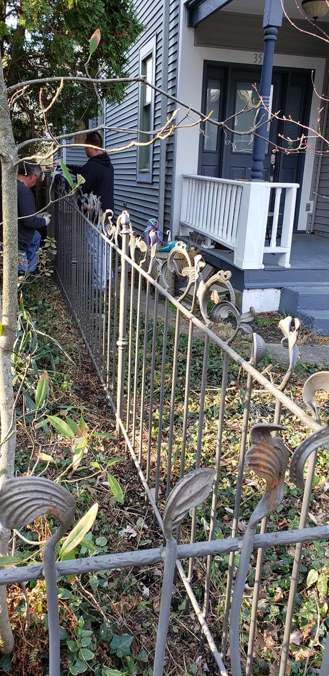 A wrought iron fence in front of a house. Two people trim a tree.