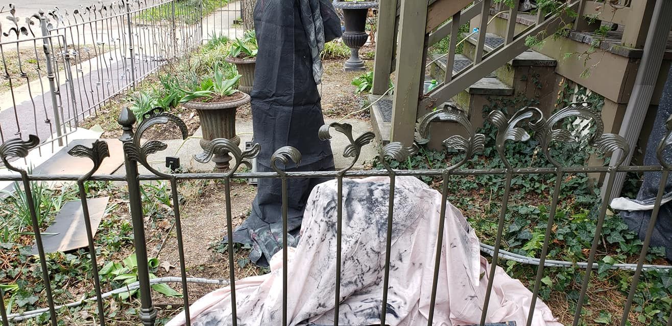 Black-clad figure behind a wrought-iron fence; garden setting with potted plants, ivy.