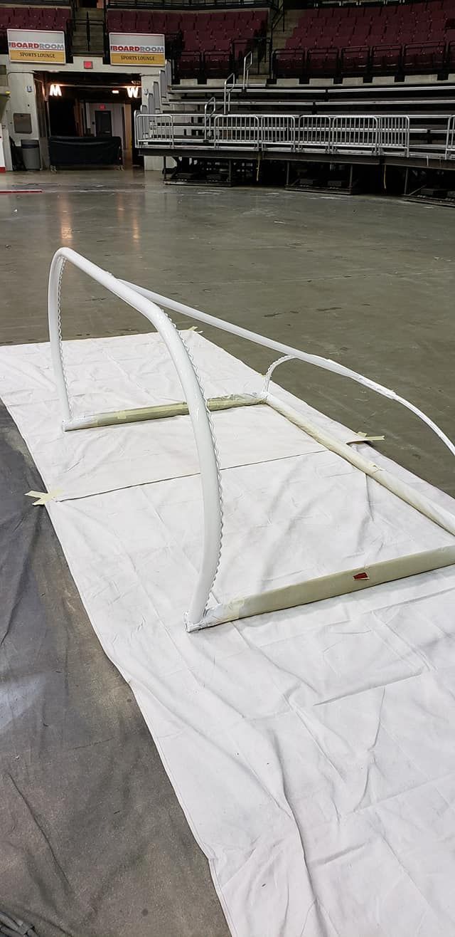 A white arched metal frame stands on a white drop cloth on the concrete floor of a large indoor arena.