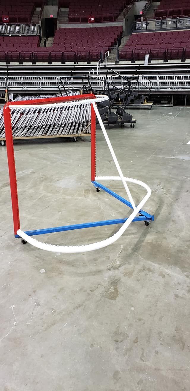 Hockey net on arena floor, red posts, blue base, white net and supports. Bleachers in background.