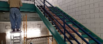 A person on a ladder working on a teal and blue staircase with white tiled walls.