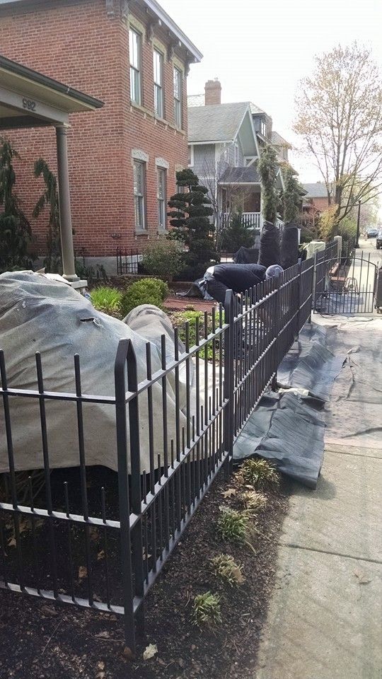 Black metal fence along a sidewalk in front of a brick house. Car partially covered with a tarp.