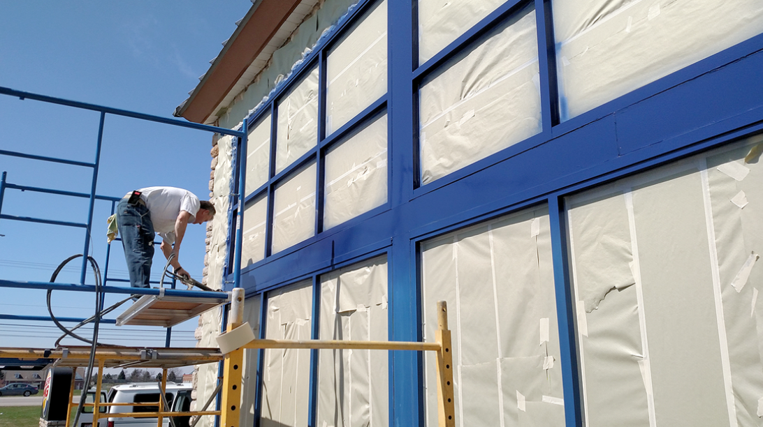 A person on scaffolding paints the blue frame of a building's windows