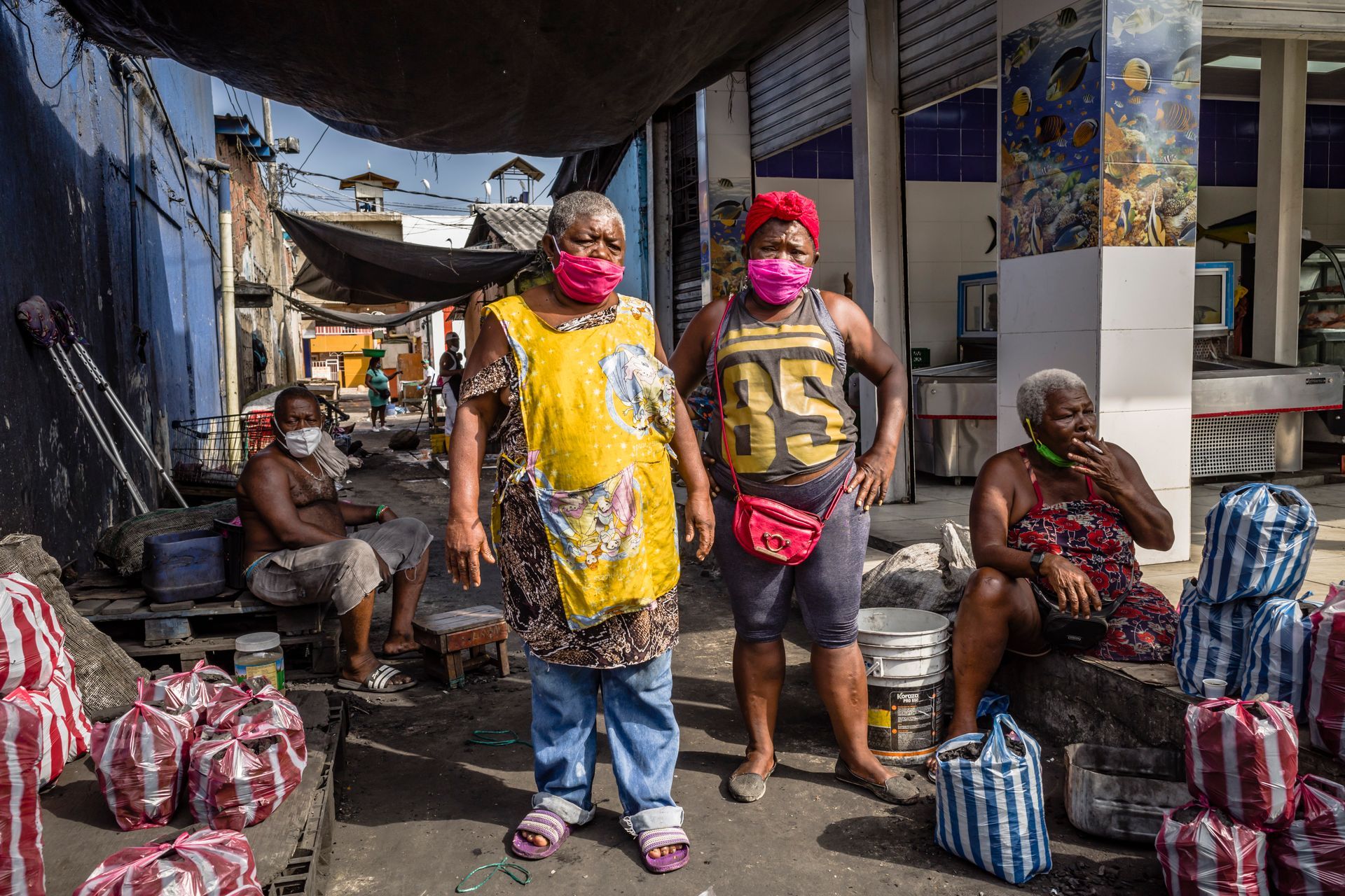 Escena de un mercado callejero con tres personas enmascaradas junto a bolsas apiladas.
