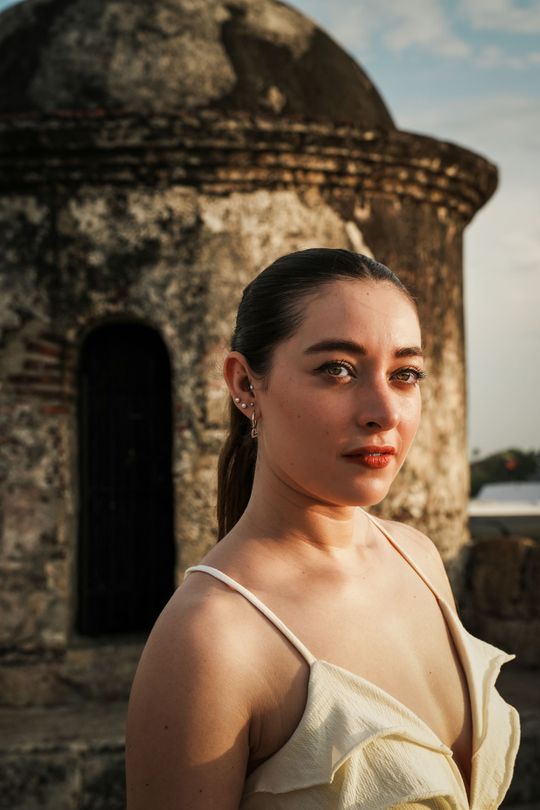 Mujer con vestido color crema de pie junto a una torre de piedra al atardecer.