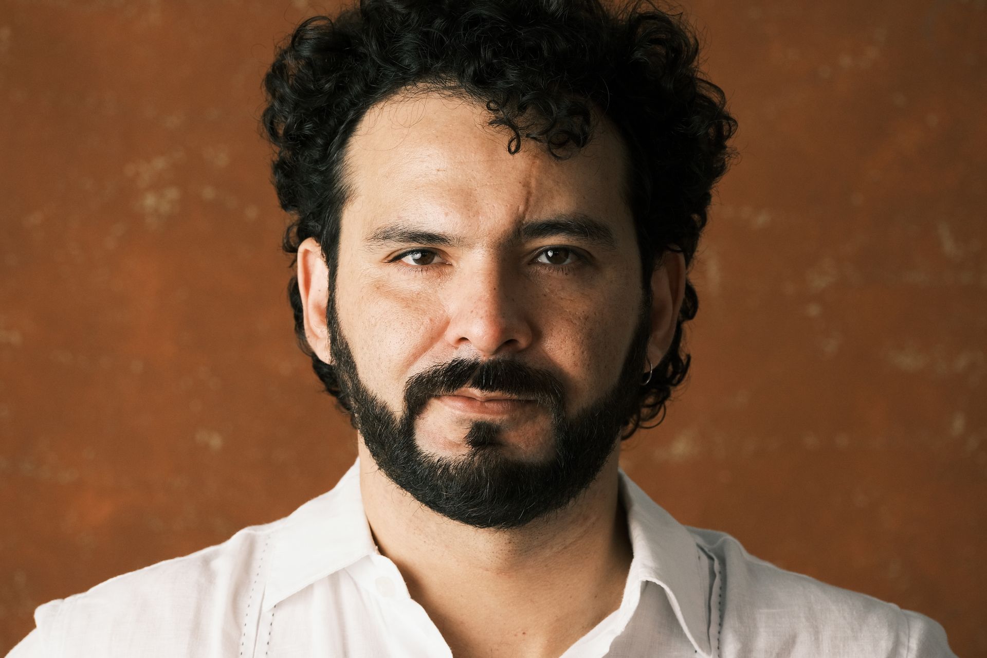 Retrato de un hombre con cabello oscuro rizado y barba, vistiendo una camisa blanca sobre un fondo marrón cálido.