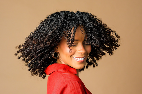 Persona sonriente con abundante cabello rizado, vestida con una blusa roja sobre un fondo beige.