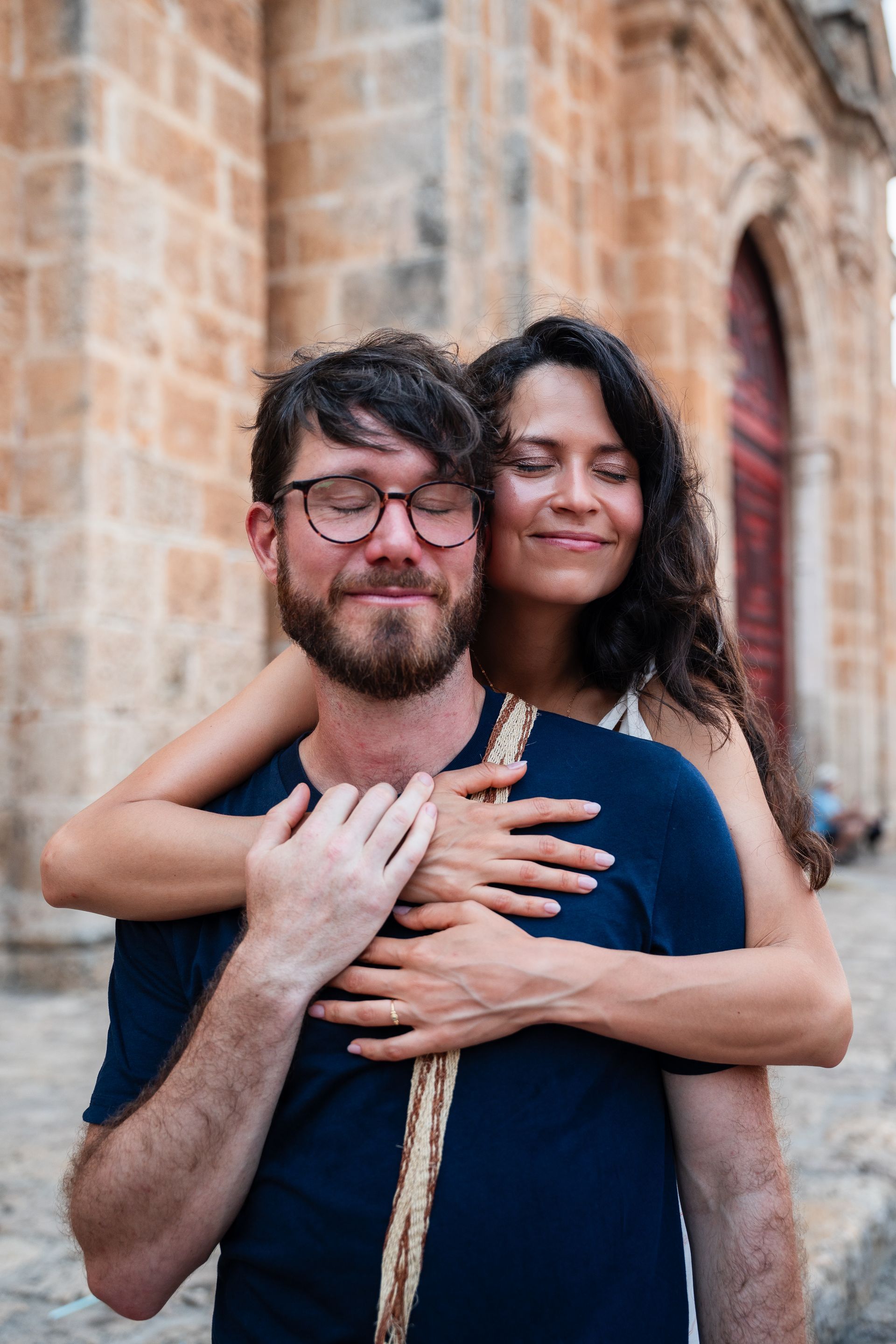 Dos personas se abrazan al aire libre junto a un edificio de piedra, ambas sonriendo con ternura.