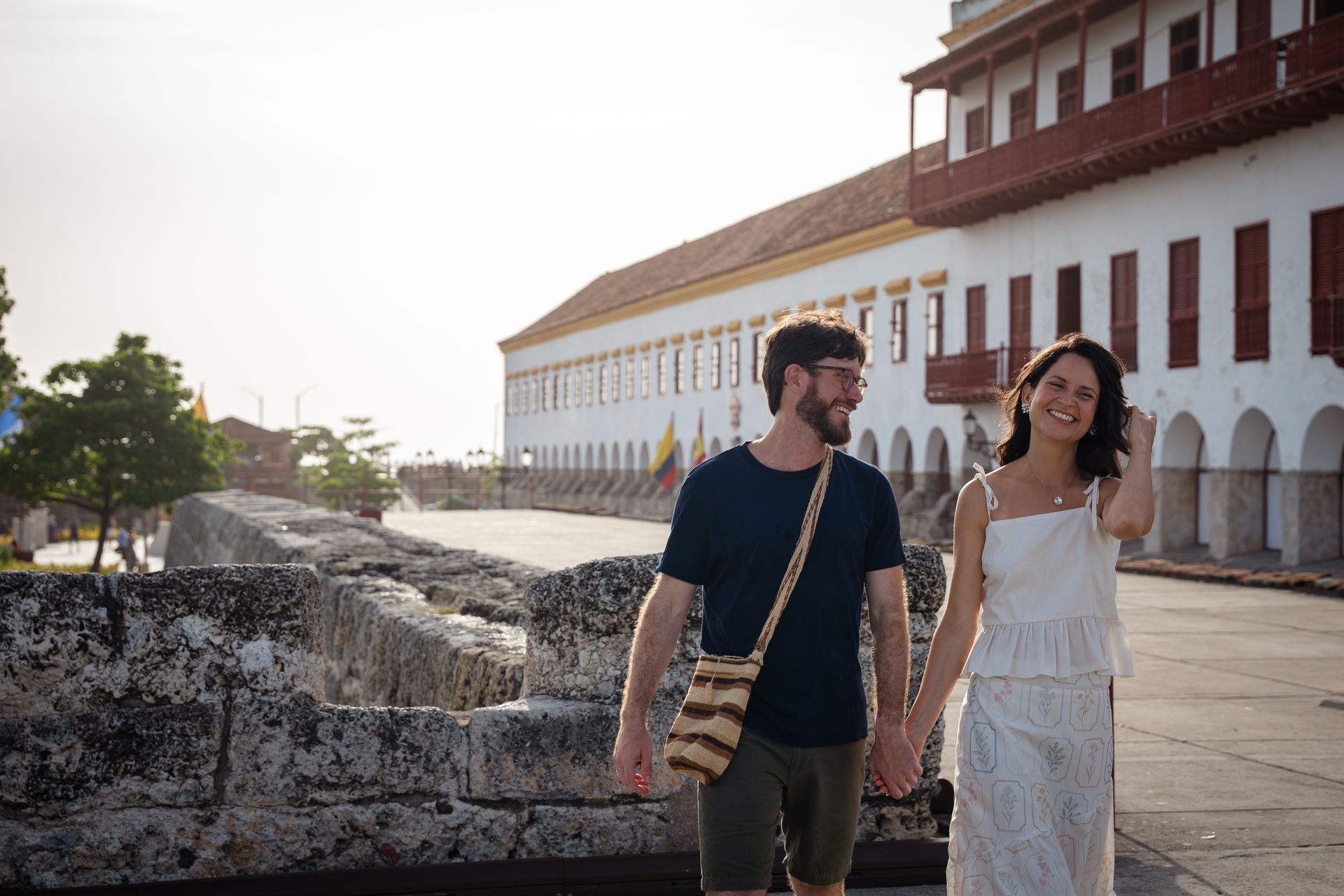 Dos personas caminan junto a un edificio blanco histórico bajo la cálida luz del sol, sonriendo y conversando.