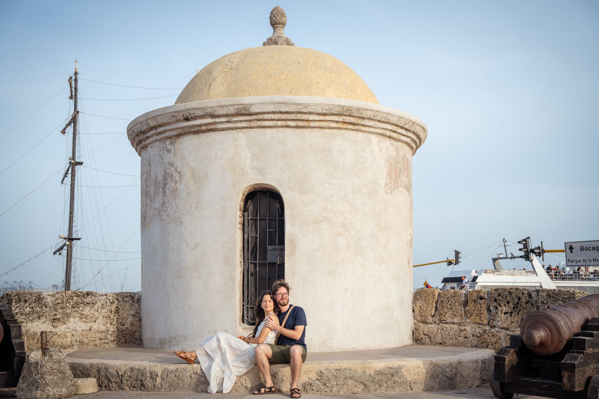 Una pareja sentada junto a una pequeña capilla blanca con cúpula, a la orilla del mar, bajo un cielo pálido.