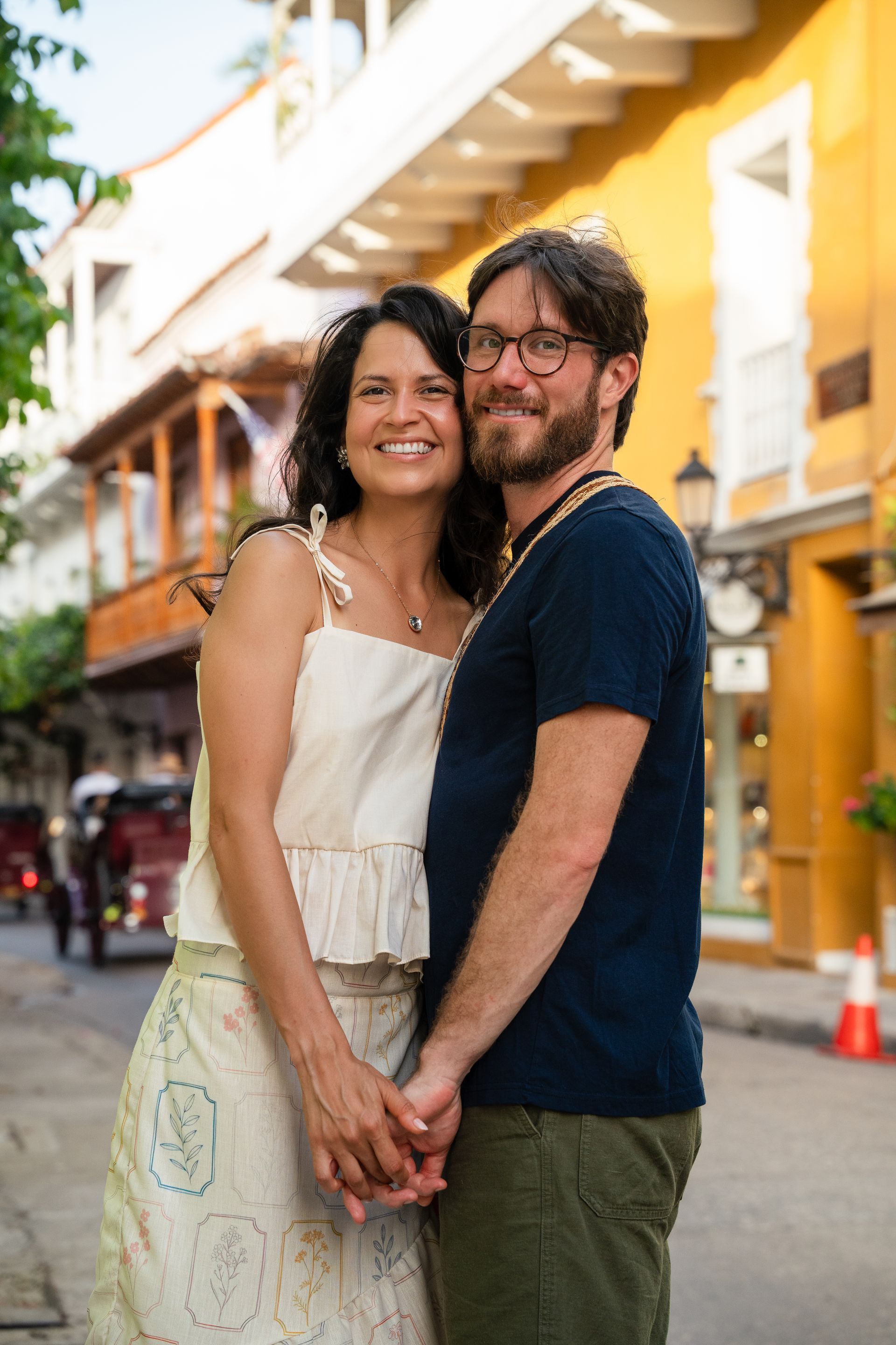 Una pareja de pie, muy juntos, en una calle colorida, sonríen a la cámara.