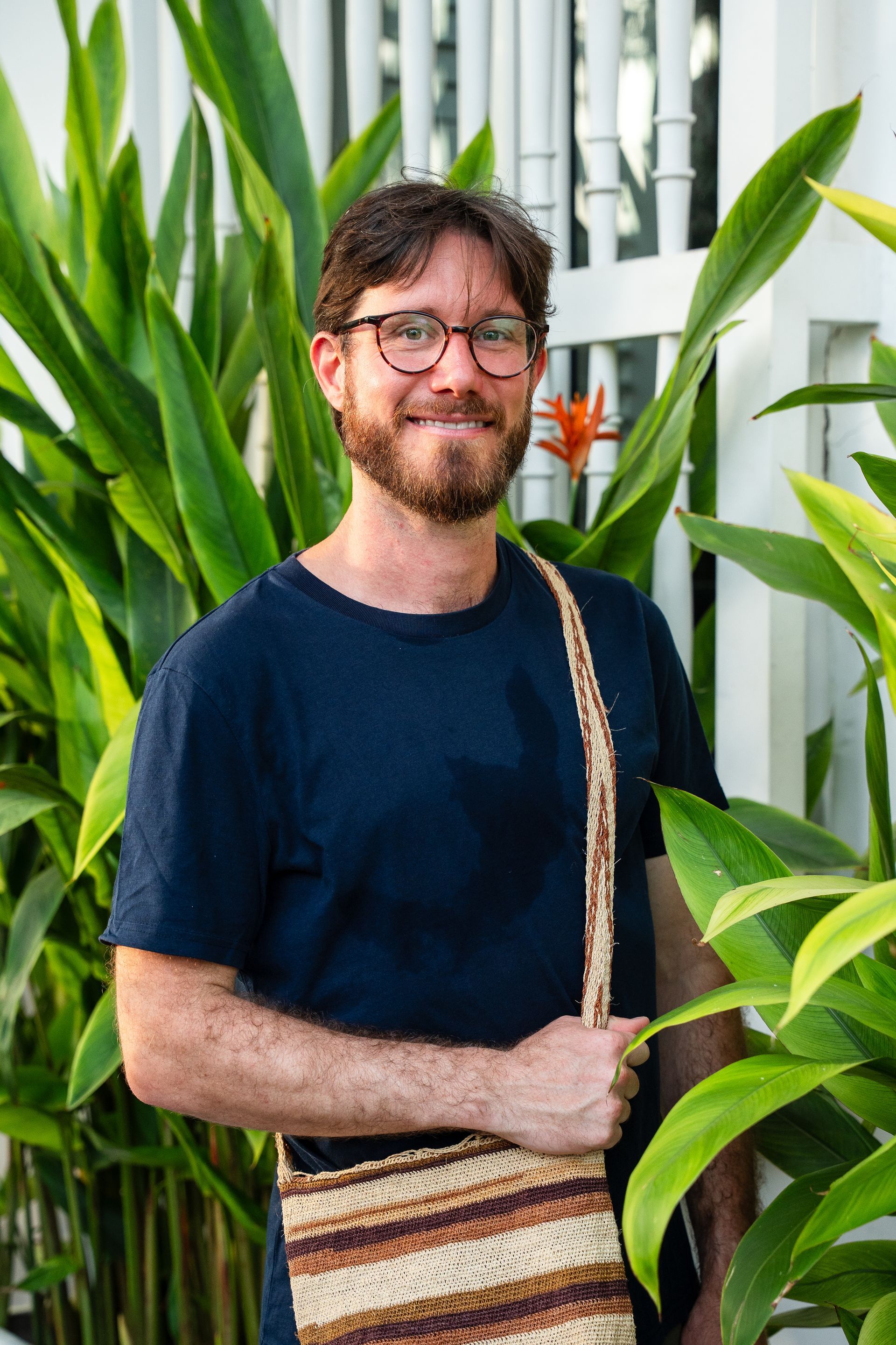 Hombre sonriente con gafas y bolso de hombro a rayas de pie entre plantas verdes altas al aire libre.
