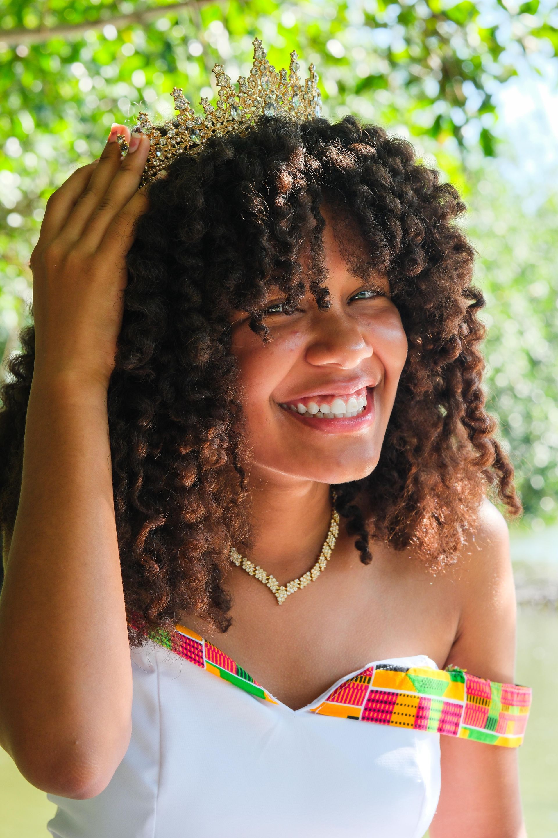 Mujer sonriente con una blusa y una corona coloridas, tocándose el cabello rizado al aire libre bajo la luz del sol.