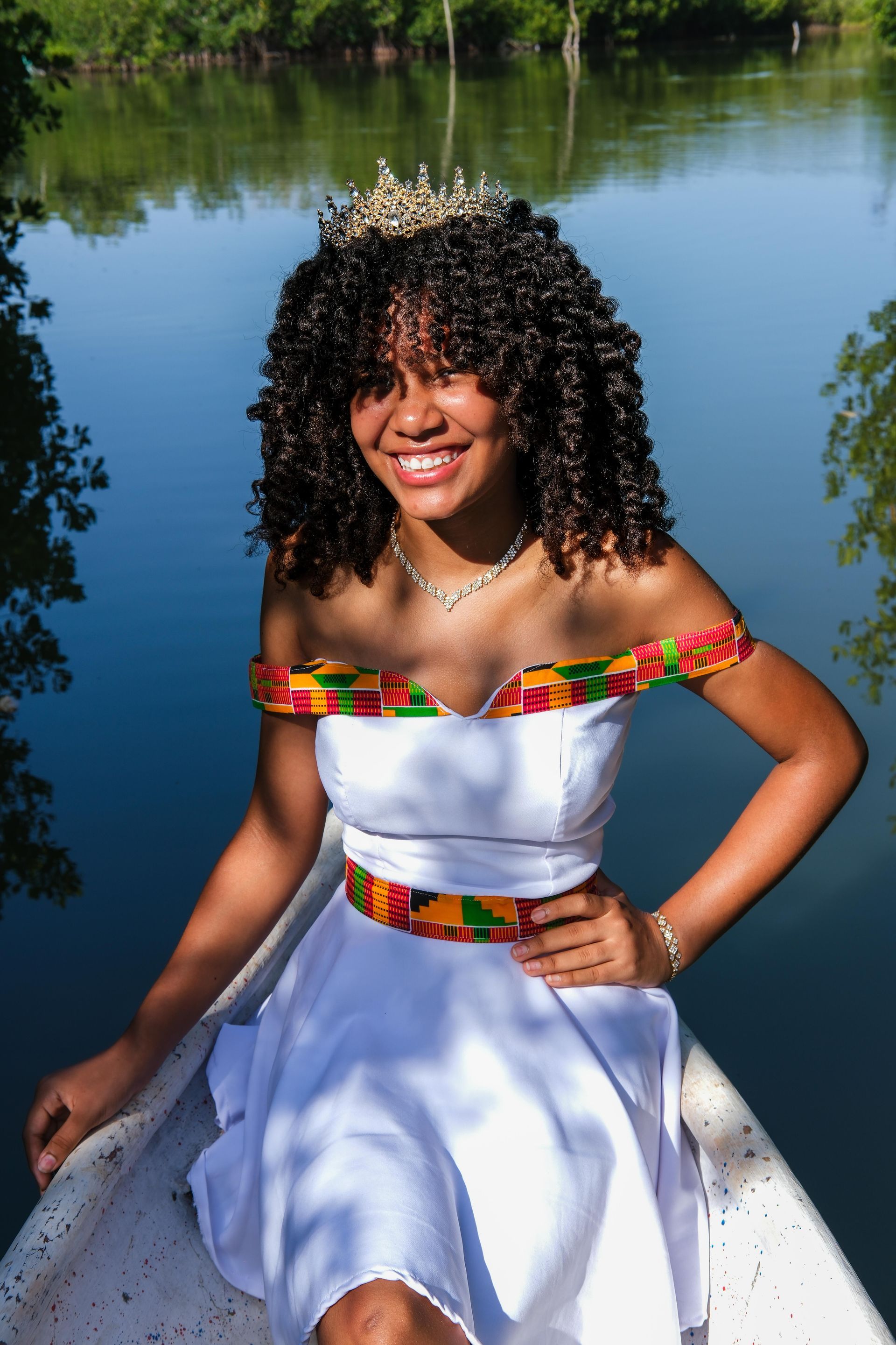 Una mujer con un vestido blanco con ribetes coloridos está sentada en un muelle junto a un lago tranquilo, sonriendo.