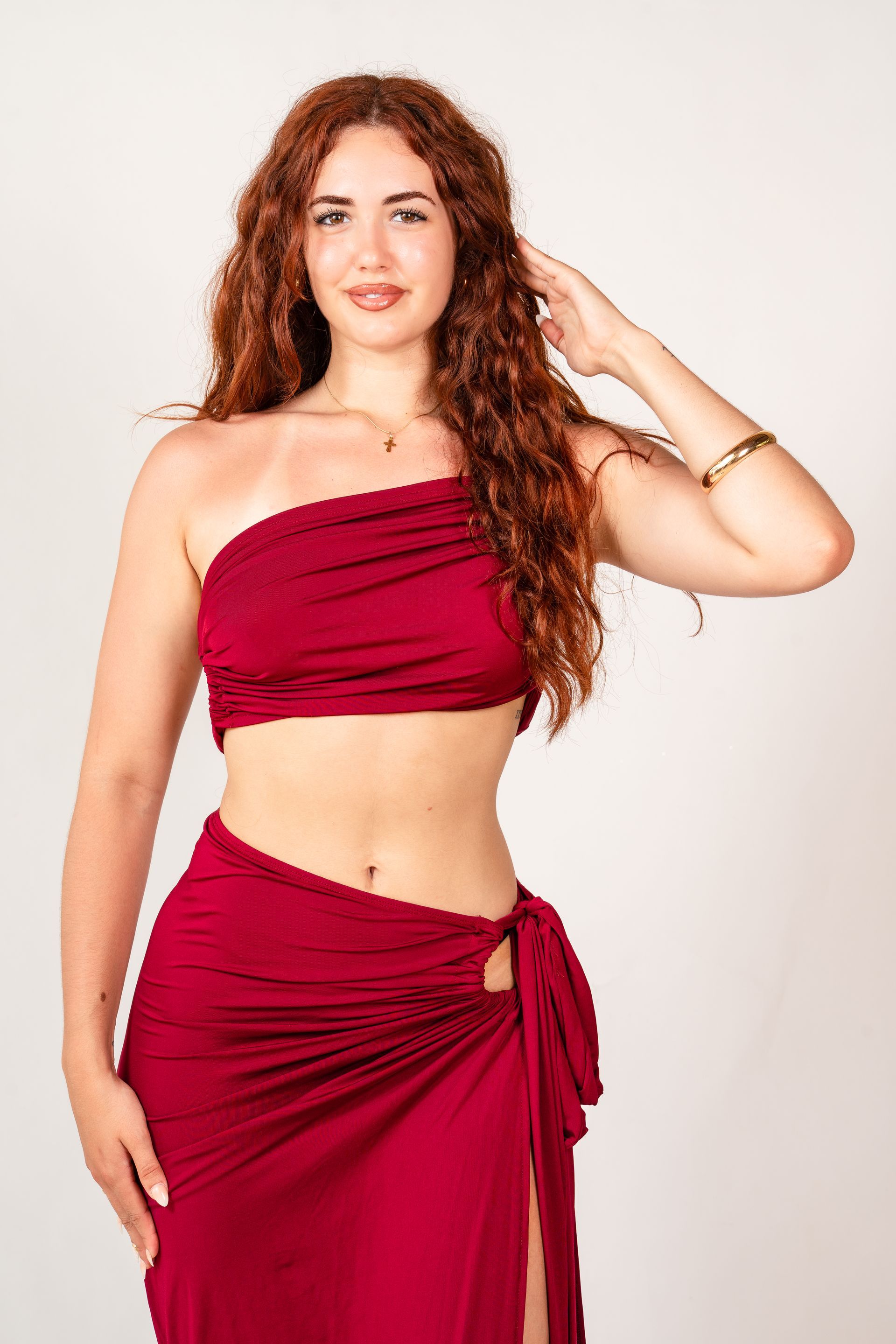 Mujer con top rojo sin tirantes y falda cruzada, de pie sobre un fondo claro, con la mano tocándose el pelo.