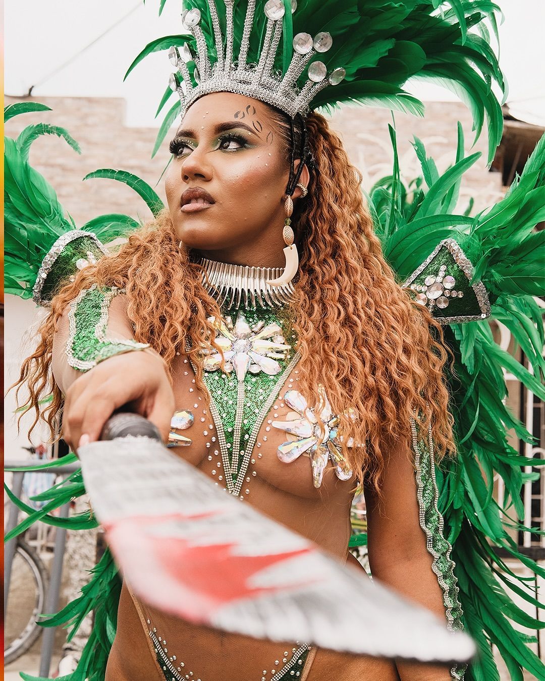 Artista de carnaval con traje de plumas verdes y tocado enjoyado, sosteniendo un abanico decorado.