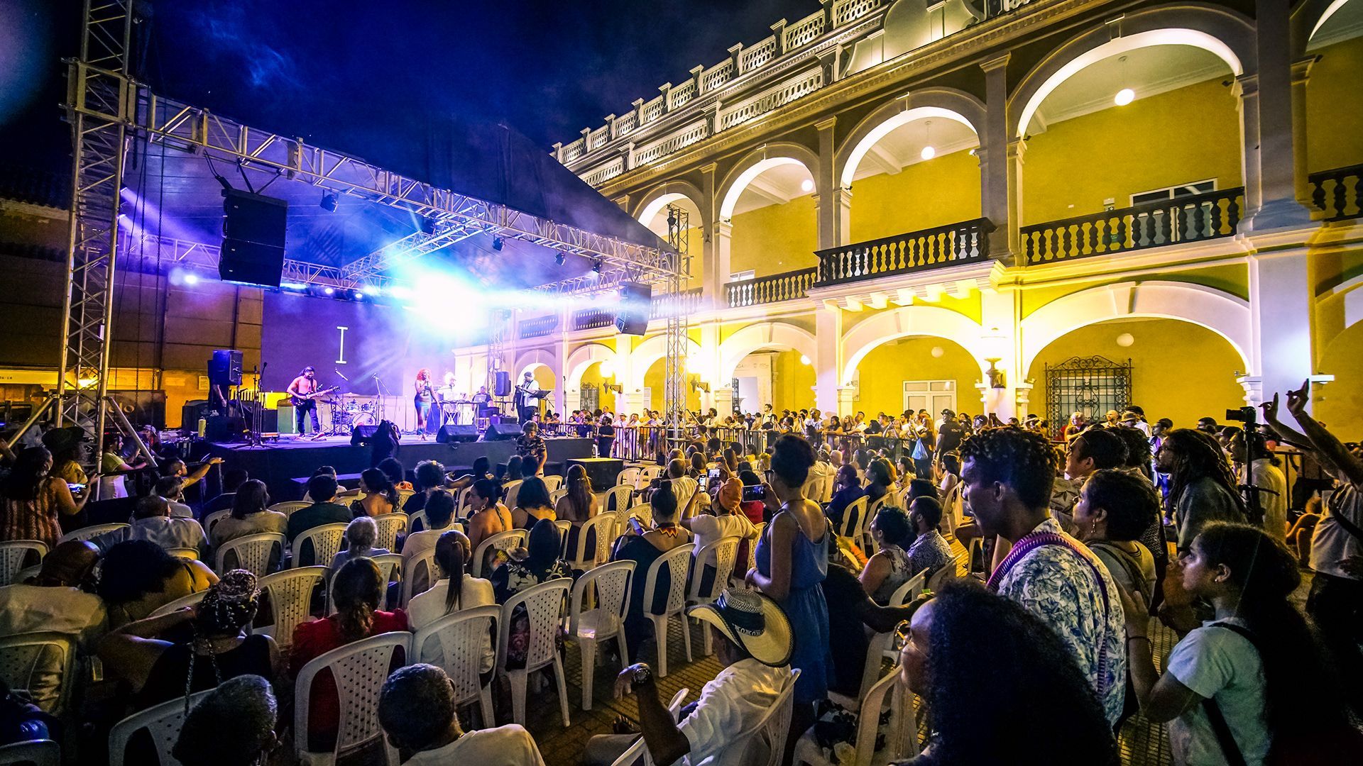 Concierto al aire libre con un escenario iluminado y una gran multitud frente a un edificio arqueado por la noche.