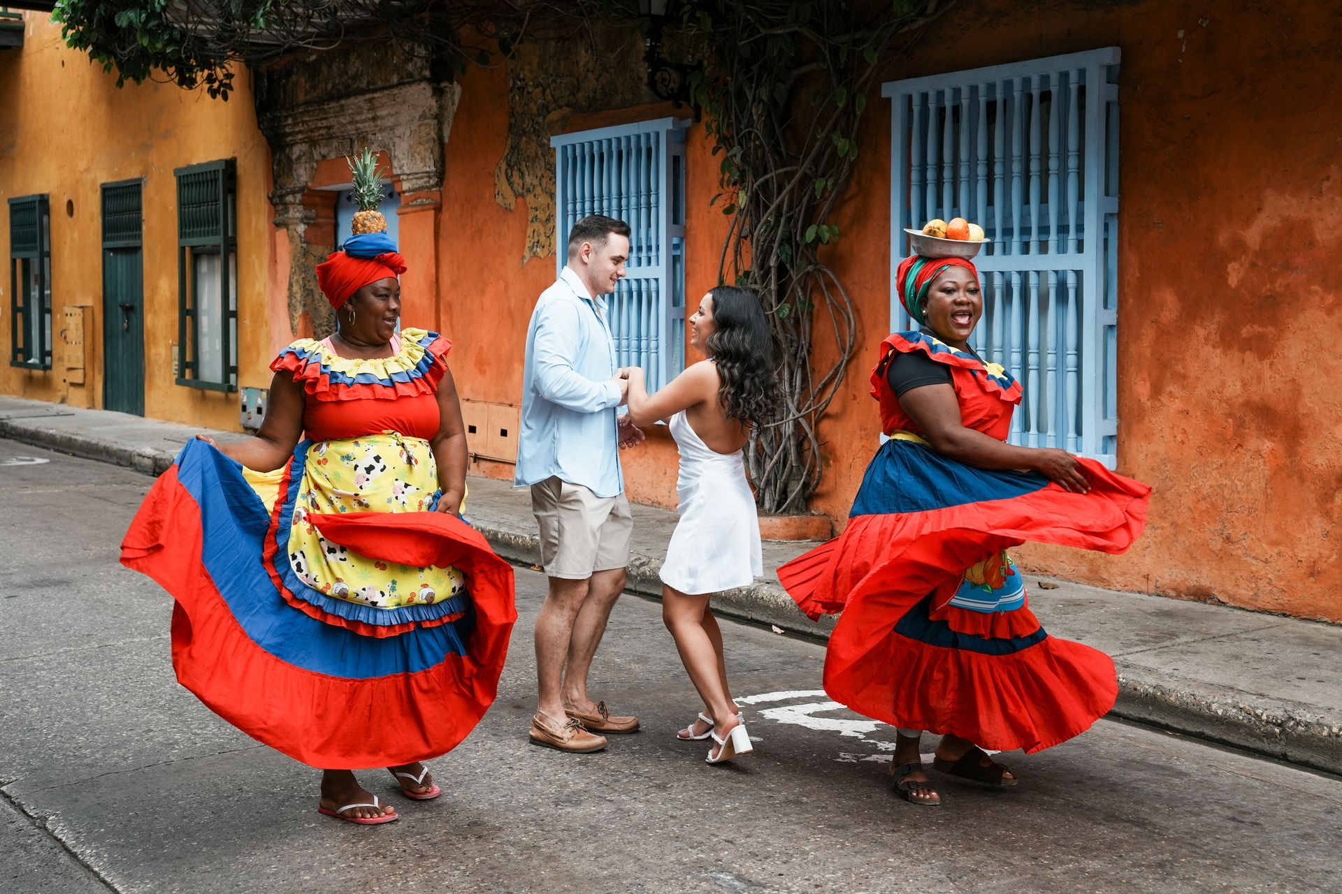 Dos bailarines con trajes rojos y azules actúan en la calle mientras una pareja camina junto a coloridos edificios.