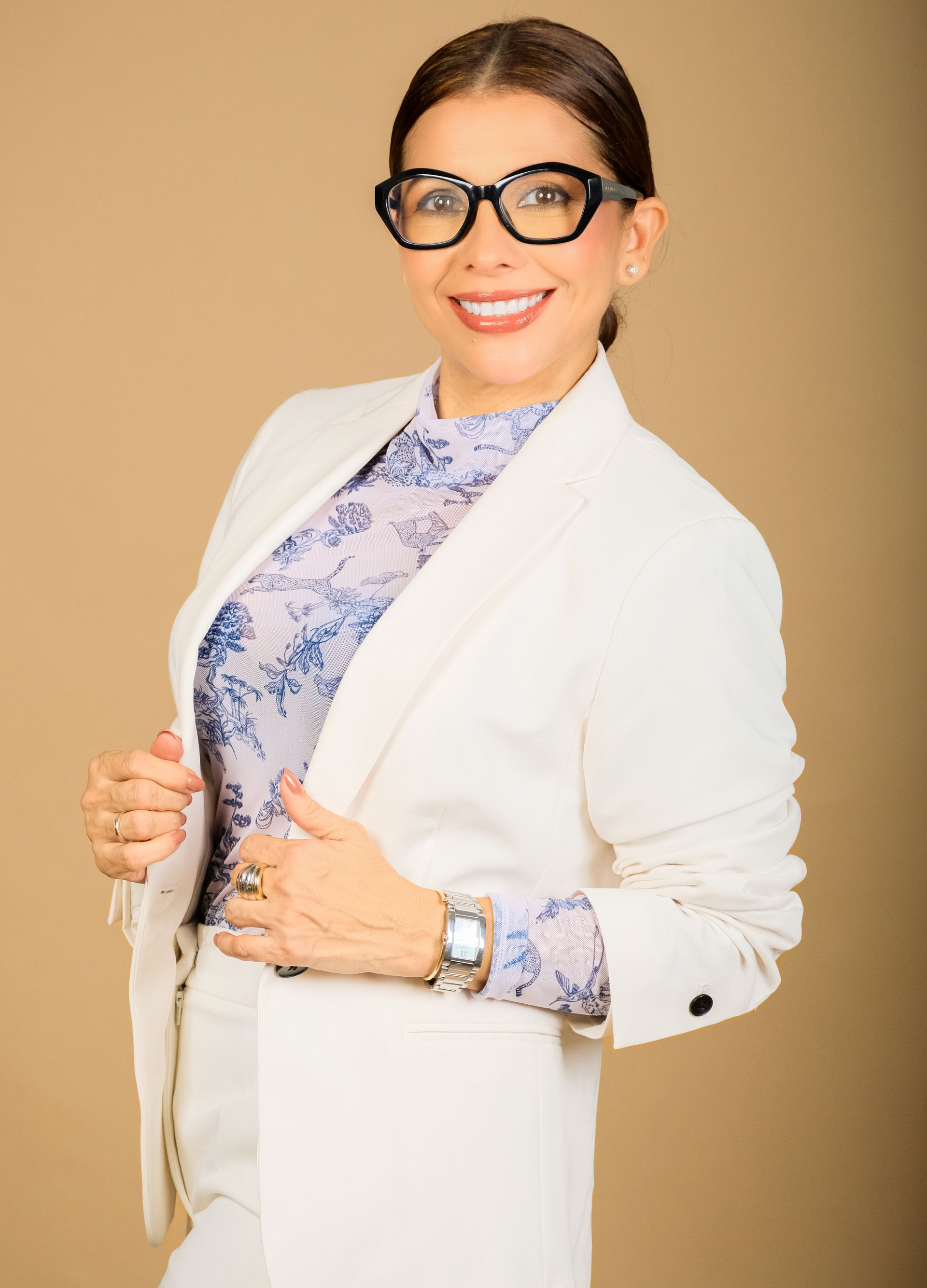 Mujer con blazer blanco y blusa estampada, sonriendo y ajustándose la chaqueta sobre un fondo beige.