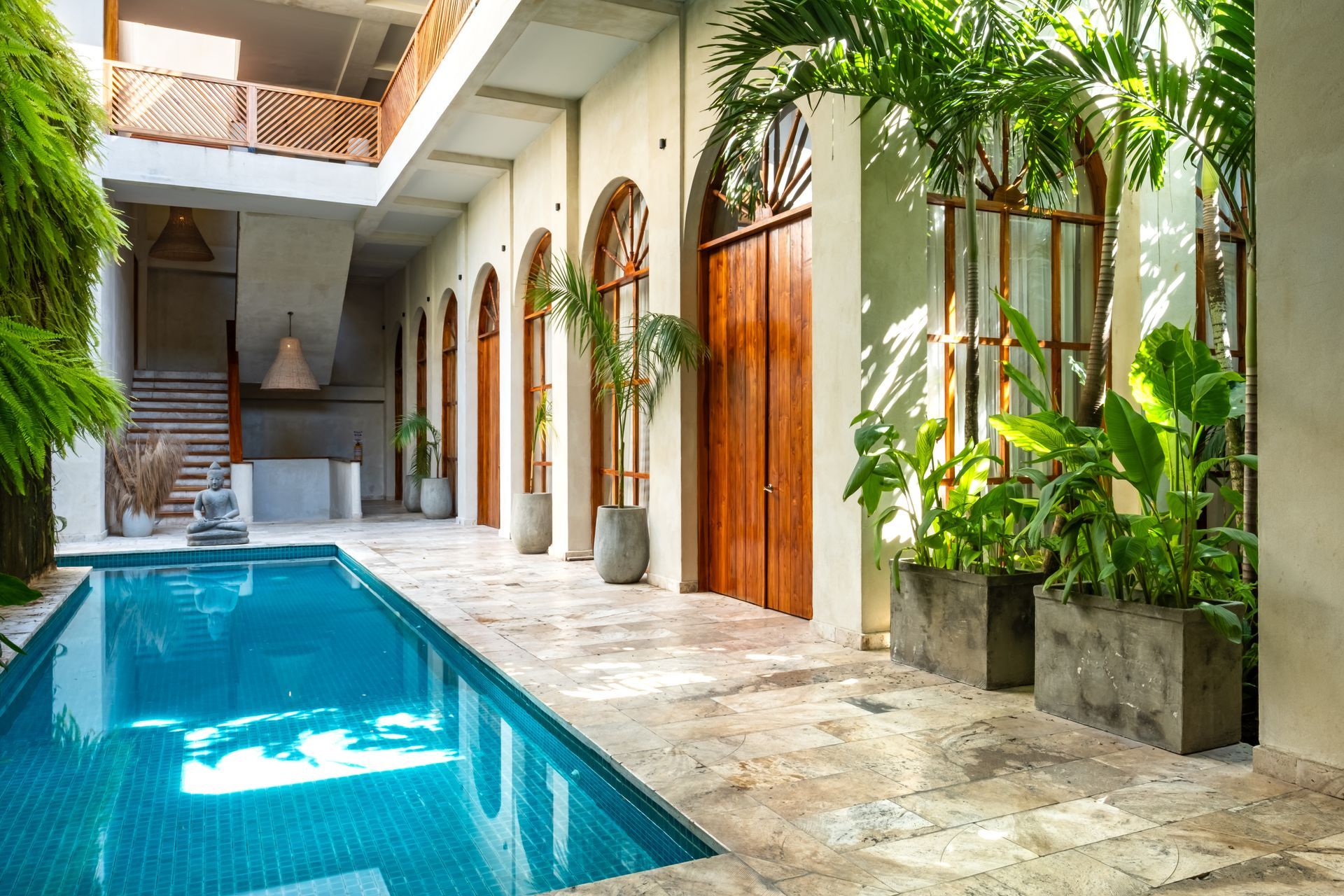 Patio con piscina turquesa, suelo de piedra, puertas de madera y exuberantes plantas tropicales bajo una claraboya.