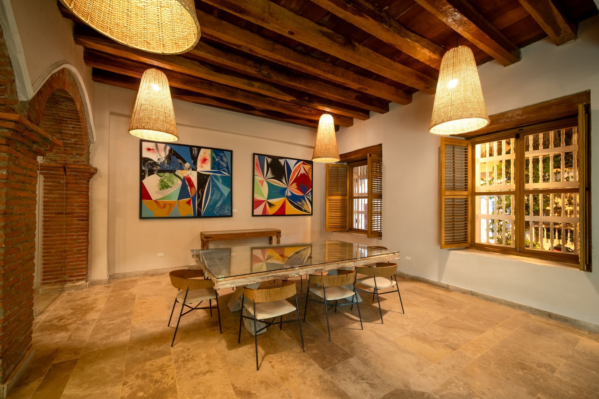 Comedor rústico con vigas de madera, lámparas colgantes, mesa de cristal, coloridos cuadros en la pared y una gran ventana.
