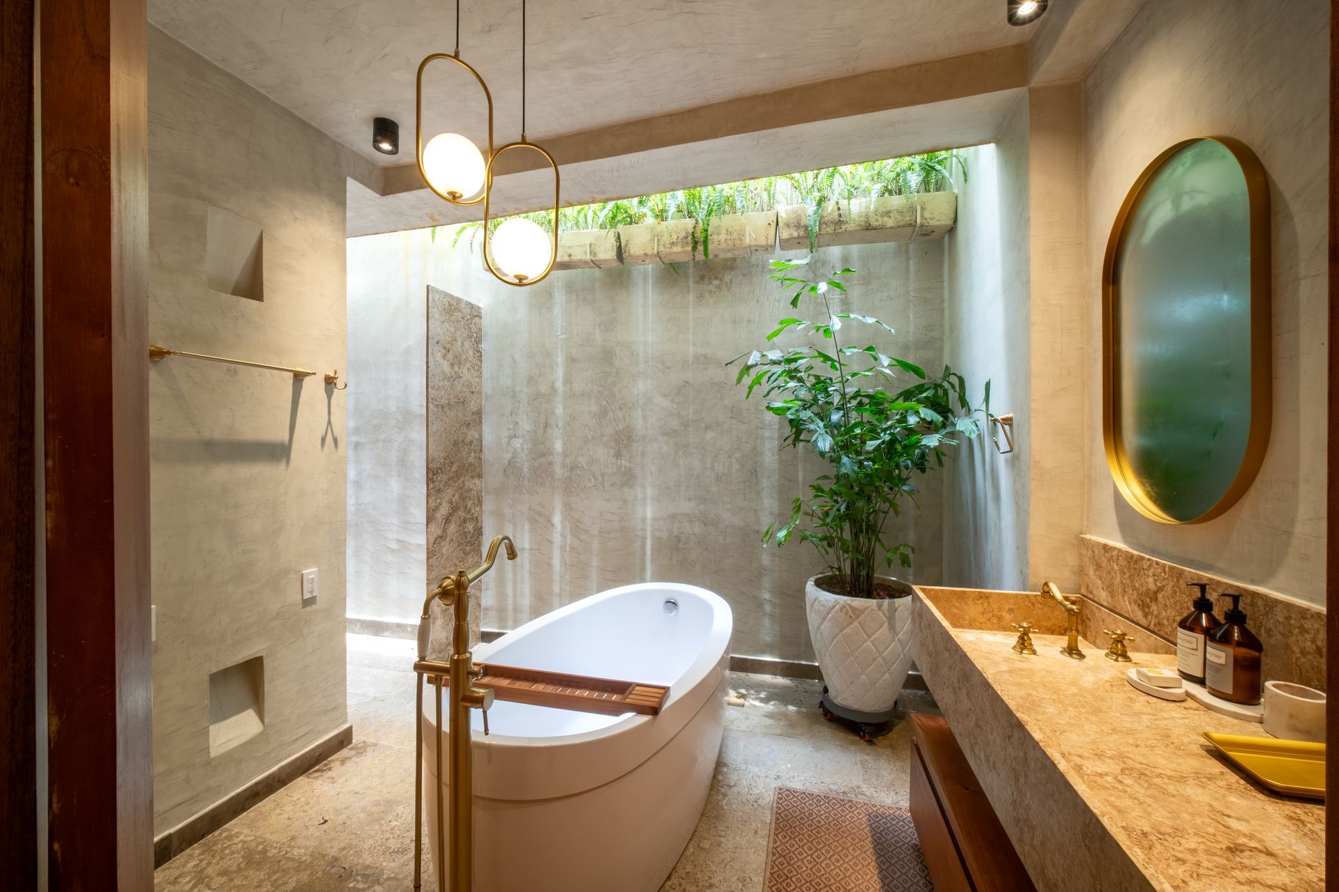 Baño moderno con bañera independiente, lavabo suspendido, espejo redondo y planta en maceta bajo una claraboya.